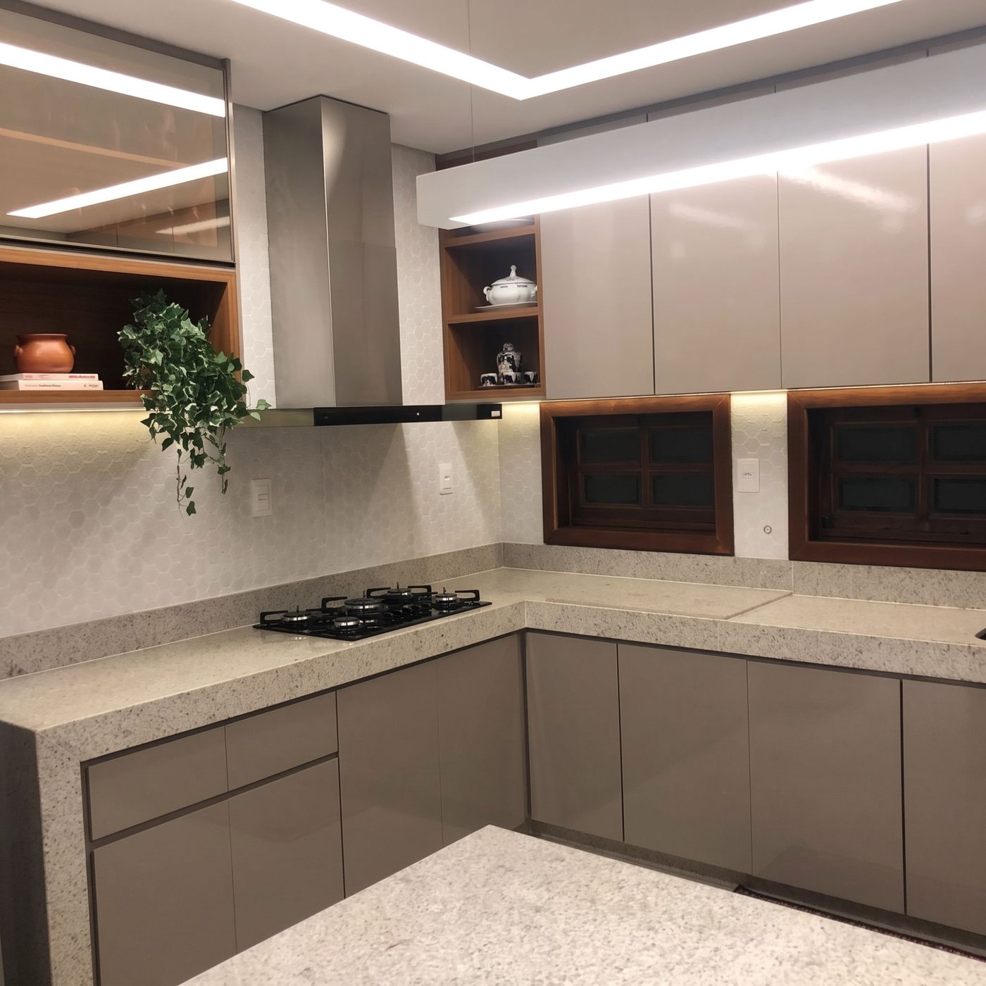 Cozinha em L em fendi, com bancada de granito, coifa de inox, nicho de madeira e sanca de LED